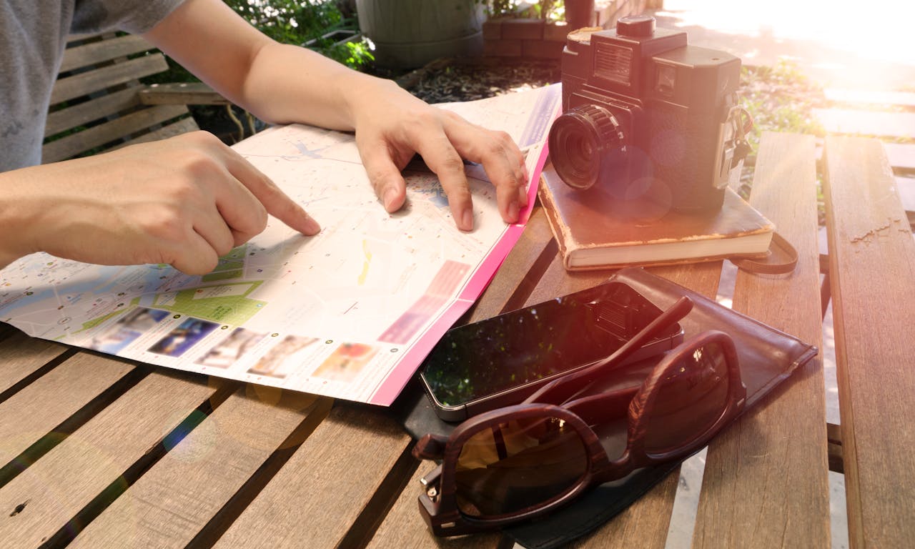 Traveler pointing at map on wooden table with camera and accessories under sunlight.